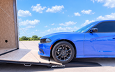 A blue sports car is positioned on a black MAXRAMPS ramp loading into a trailer outdoors under a bright blue sky with clouds.