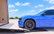 A blue sports car is positioned on a black MAXRAMPS ramp loading into a trailer outdoors under a bright blue sky with clouds.