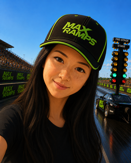 A woman wearing a black and neon green MAX RAMPS baseball cap takes a selfie on a racetrack with a drag race car and starting lights in the background.