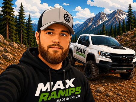 A man in a black hoodie with 'MAX RAMPS MADE IN THE USA' and a gray-and-black cap poses in front of a white Chevrolet Colorado pickup truck on a mountain trail. The scene is set against a backdrop of pine trees and snow-capped mountains.