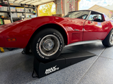 A red classic Corvette parked in a garage on black MaxRamps drive-up ramps, with the front wheel raised. The car features a glossy red paint job, chrome wheels, and a visible Winston tire.