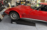 A red classic Corvette parked in a garage on a black MaxRamps drive-up ramp, with shelves and tools visible in the background.