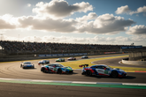 A group of racing cars navigates a track under a cloudy sky, with grandstands filled with spectators in the background.